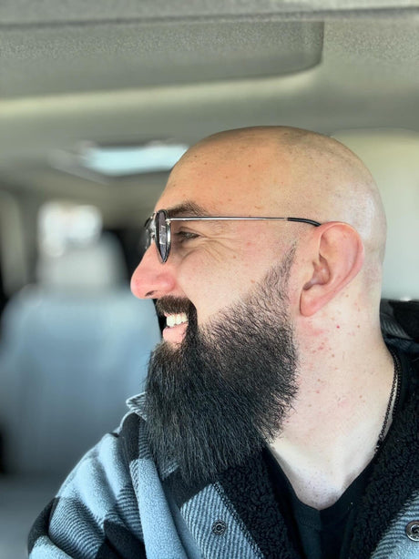 Profile of a smiling man with a bald head and a beard, wearing glasses, in the passenger seat of a car using Mountaineer Brand Products' Cleanse & Protect Combo.