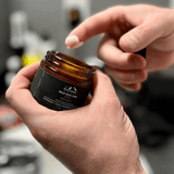 A person is holding a jar of Timber Beard Balm by Mountaineer Brand Products, an all-natural cream.