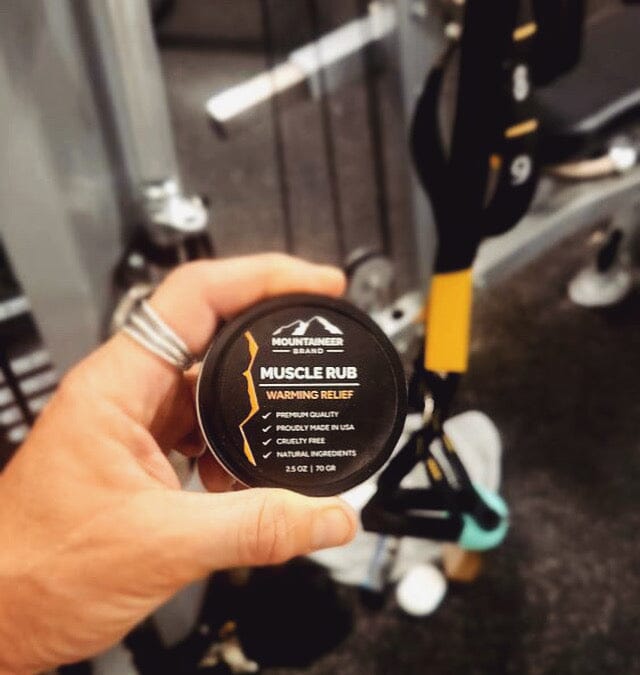 Hand holding a container of Mountaineer Brand Products Muscle Rub in a gym setting.