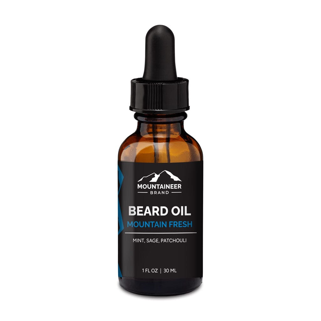 A glass bottle of Mountaineer Brand Products Natural Beard Oil labeled "mountain fresh" with mint, sage, and patchouli essential oils, featuring a dropper cap, isolated on a white background.