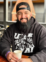 A smiling man with a beard wearing a cap and a graphic hoodie sits holding a cup at a table, showcasing his well-groomed beard from the "Mountaineer Brand Products Bald and Bearded Kit.