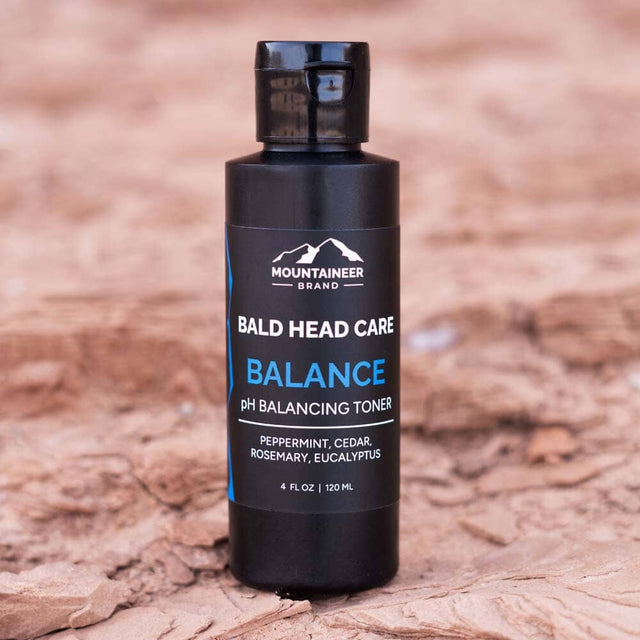 A bottle of Mountaineer Brand Products Bald Head Shine Away PH Balancing Toner with natural ingredients like peppermint, cedar, rosemary, and eucalyptus, displayed on a textured surface.