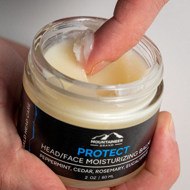 A person's finger touching a jar of Mountaineer Brand Products Bald Head Protect (Moisturizer) balm.