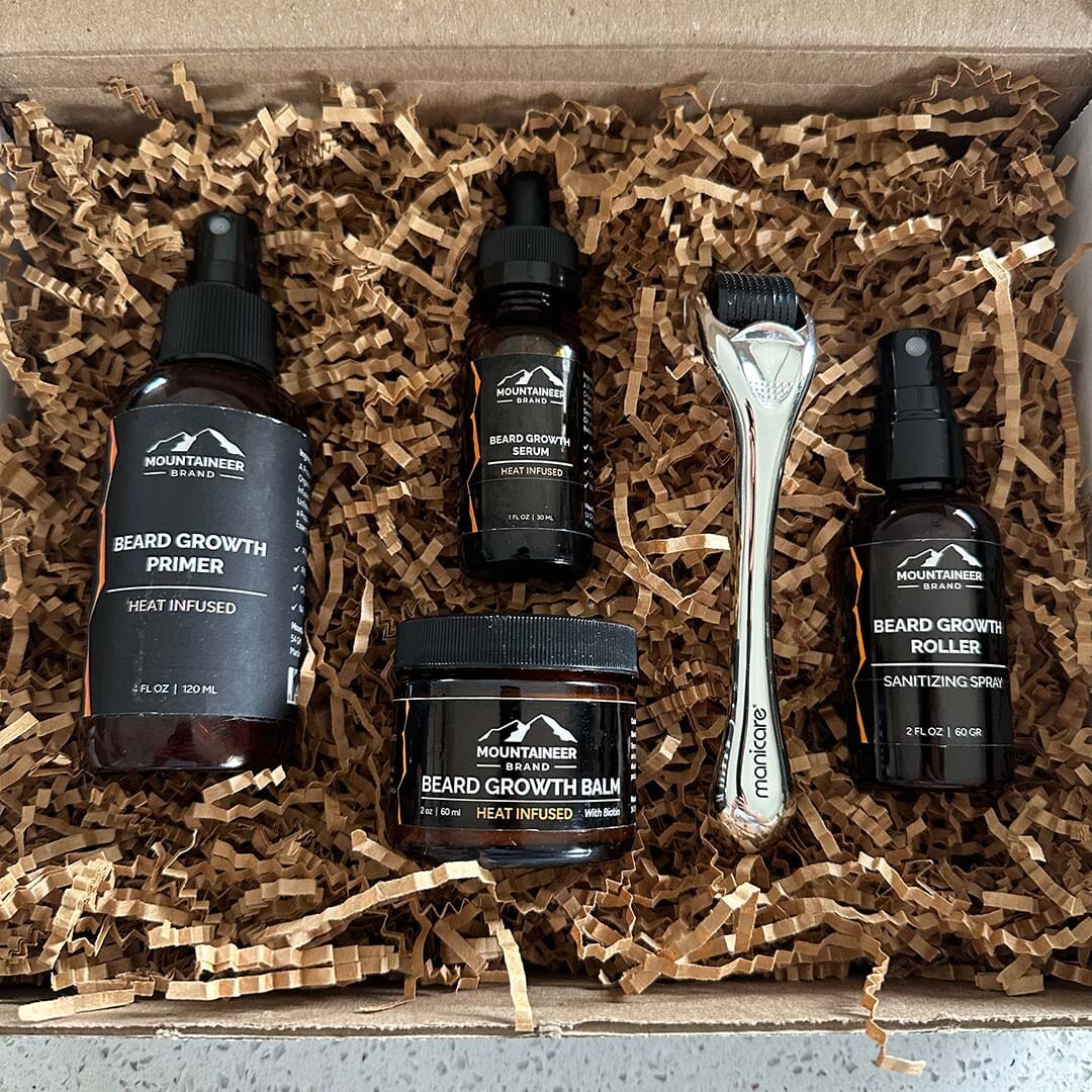 A selection of Essential Beard Growth System products including Beard Growth Serum, balms, and a razor, displayed inside a box with shredded paper filling by Mountaineer Brand Products.