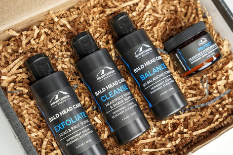 Four bottles of Mountaineer Brand Products' The Essential Bald Head Care System arranged neatly in a box filled with shredded paper, containing essential oils and natural ingredients.