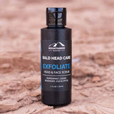A bottle of Mountaineer Brand Products Bald Head Exfoliater on a dry, cracked ground.