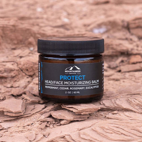 A jar of Mountaineer Brand Products Bald Head Protect moisturizer on a dry, cracked earth background.