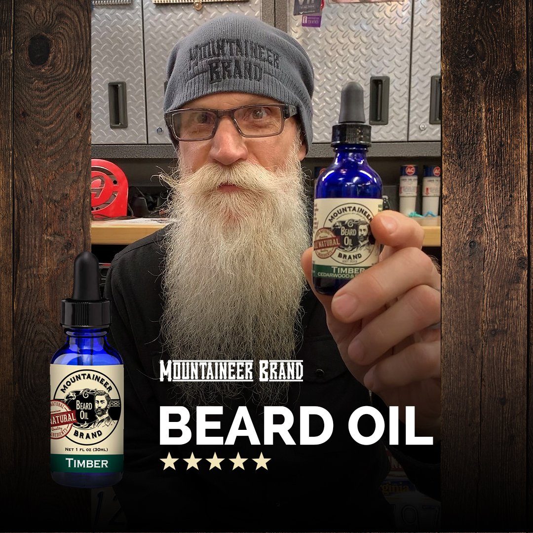 Man with a long beard wearing glasses and a beanie, holding a bottle of Mountaineer Brand Products Natural Beard Oil in a workshop setting.