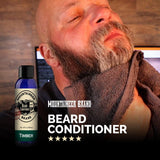 A bald man with a beard and closed eyes, feeling his soft beard, next to a bottle of Mountaineer Brand Products' Natural Beard Conditioner with five-star reviews.
